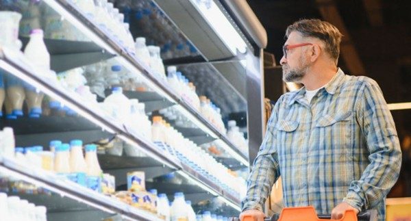 A man with glasses and a shopping cart observes dairy products on a refrigerated shelf in a supermarket, illustrating in-store shopper behavior and product engagement.